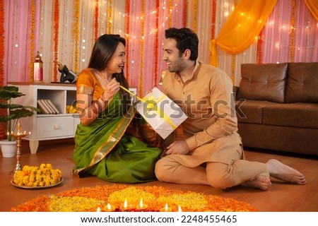 Diwali Celebration India - Happy Indian wife opening a gift given by her husband during festival . Royalty free image of a couple sitting together and making rangoli - wife opening the gift given ... Royalty-Free Stock Photo #2248455465