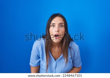 Young brunette woman standing over blue background afraid and shocked with surprise and amazed expression, fear and excited face. 