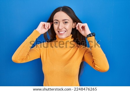 Young brunette woman standing over blue background smiling pulling ears with fingers, funny gesture. audition problem 