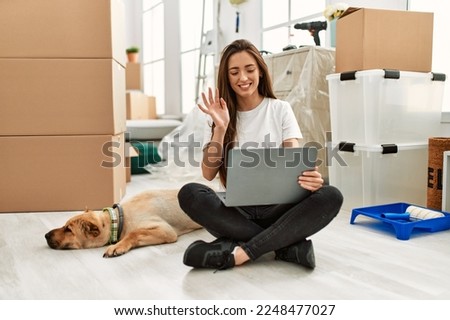 Young hispanic woman having video call sitting on floor with dog at new home