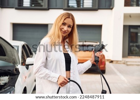 With charging cable in hands. Young woman in white clothes is with her electric car at daytime.