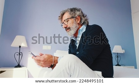 Stylish good looking mature man is sitting on bed and using smartphone