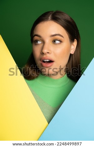 Shocked teen girl with visage looking away near colorful paper isolated on green Royalty-Free Stock Photo #2248499897