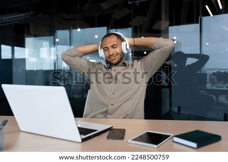 Successful young african american businessman relaxing in office, man in shirt and headphones listening to music online radio and podcasts, sitting at desk using laptop at work. Royalty-Free Stock Photo #2248500759