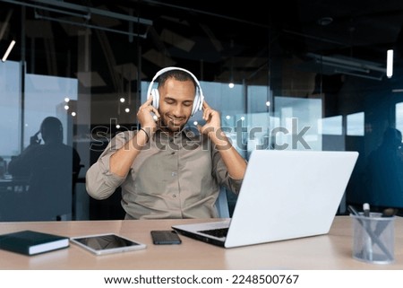 Successful young african american businessman relaxing in office, man in shirt and headphones listening to music online radio and podcasts, sitting at desk using laptop at work. Royalty-Free Stock Photo #2248500767