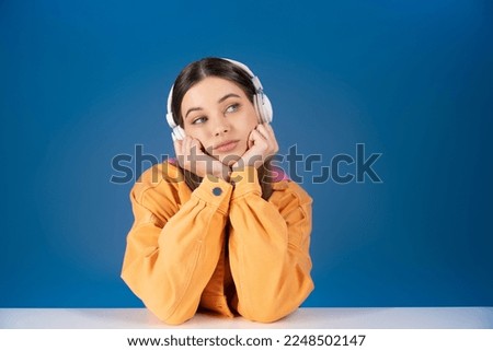 Dreamy brunette teen girl in headphones looking away near table isolated on blue
