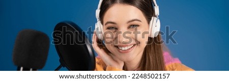 Teen podcaster in headphones smiling at camera near studio microphone isolated on blue, banner