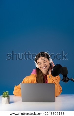 Teen podcaster in headphones looking at laptop near studio microphone isolated on blue