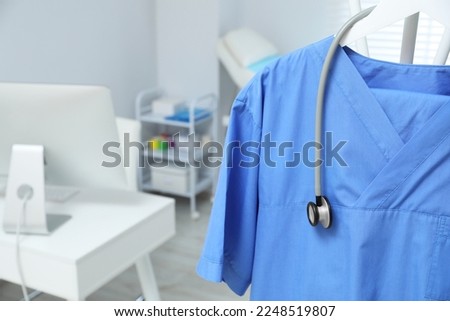 Blue medical uniform and stethoscope hanging on rack in clinic, closeup. Space for text