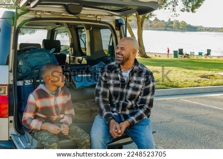 Landscape photo of father and son laughing and sitting in back of car getting ready to unpack for camping.