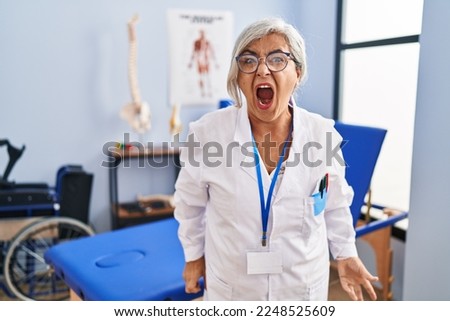 Middle age woman with grey hair working at pain recovery clinic angry and mad screaming frustrated and furious, shouting with anger. rage and aggressive concept. 