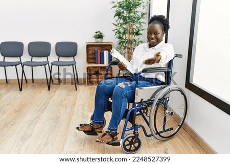 Young black woman sitting on wheelchair at waiting room smiling cheerful offering palm hand giving assistance and acceptance. 