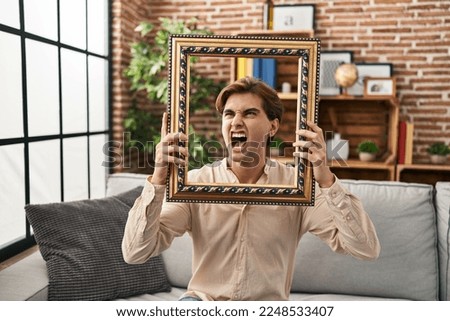 Young man holding empty frame angry and mad screaming frustrated and furious, shouting with anger. rage and aggressive concept. 