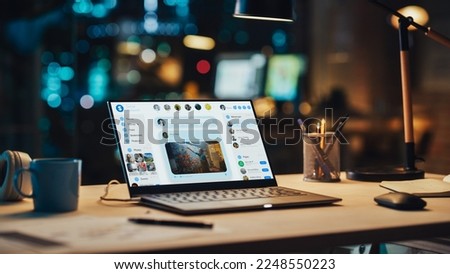 Laptop Standing on a Desk with a Social Media Platform Displayed on Screen. Table with Computer, Coffee Mug, Headphones and Notebook in Creative Office at Night. Royalty-Free Stock Photo #2248550223