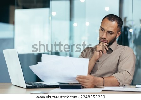 Pensive serious businessman reading financial report, hispanic businessman holding document in hands looking disappointed, working inside modern office with laptop behind paper work. Royalty-Free Stock Photo #2248555239