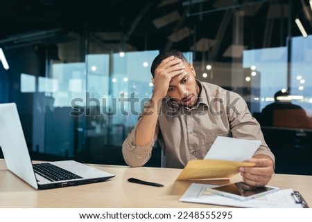 Pensive businessman inside office reading notification letter, man received envelope thinking about decision sitting at table and laptop inside office, bad news message. Royalty-Free Stock Photo #2248555295