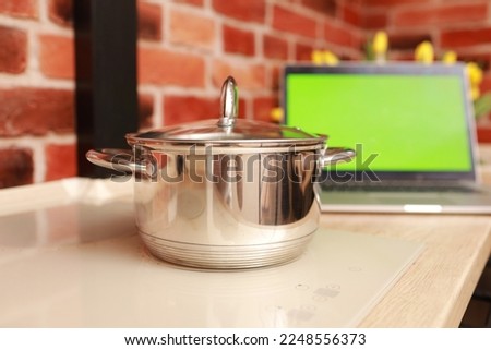 Steel saucepan on white stylish induction stove and laptop with green screen with chrome key at kitchen on brick wall background. Cooking and vlogging concept.