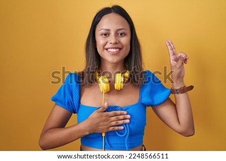 Hispanic young woman standing over yellow background smiling swearing with hand on chest and fingers up, making a loyalty promise oath 