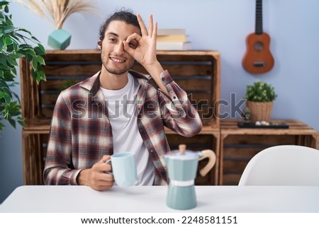 Young hispanic man drinking coffee from french coffee maker smiling happy doing ok sign with hand on eye looking through fingers 