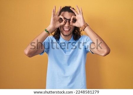Young hispanic man standing over yellow background doing ok gesture like binoculars sticking tongue out, eyes looking through fingers. crazy expression. 