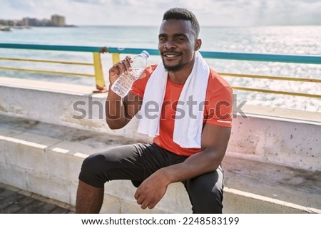 Young african american man wearing sportswear holding water bottle at seaside Royalty-Free Stock Photo #2248583199