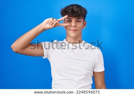 Hispanic teenager standing over blue background doing peace symbol with fingers over face, smiling cheerful showing victory 