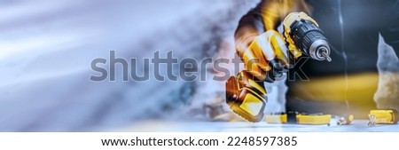Man worker holds an electric screwdriver in his hands close-up against the background of a construction tool and a concrete wall. Long banner with glow effects Royalty-Free Stock Photo #2248597385