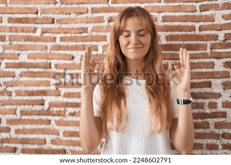 Young caucasian woman standing over bricks wall relaxed and smiling with eyes closed doing meditation gesture with fingers. yoga concept. 