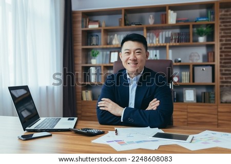 Portrait of a young handsome Asian man, businessman, boss in a suit, sitting in a chair at a table in the office, working with a laptop and documents in the office, looking at the camera. Crossed his Royalty-Free Stock Photo #2248608083