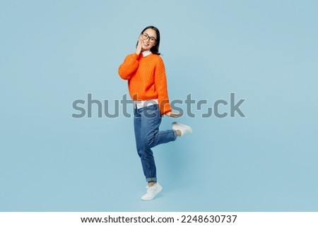 Full body side view young woman of Asian ethnicity wear orange sweater glasses look camera hold face raise up leg isolated on plain pastel light blue cyan background studio. People lifestyle concept Royalty-Free Stock Photo #2248630737