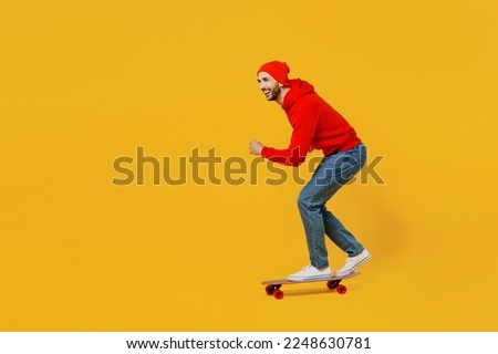 Full body side profile view smiling happy young caucasian man wear red hoody hat riding skateboard pennyboard isolated on plain yellow color background studio portrait. People urban lifestyle concept