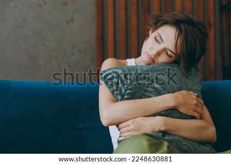 Young frustrated unhappy sad european woman wears white tank shirt hold pillow look aside sit on blue sofa couch stay at home flat spend time in living room indoors grey wall. People lounge concept Royalty-Free Stock Photo #2248630881