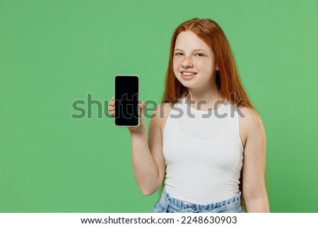 Little redhead kid girl 12-13 years old wearing white tank shirt hold in hand use mobile cell phone with blank screen workspace area isolated on plain green background. Childhood lifestyle concept.