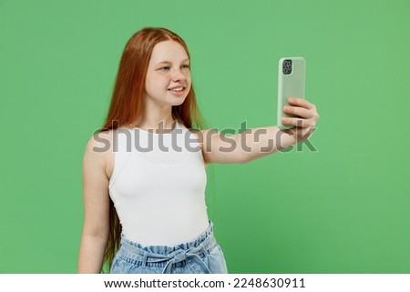 Little redhead kid girl 12-13 years old wearing white tank shirt doing selfie shot on mobile cell phone post photo on social network isolated on plain green background. Childhood lifestyle concept.