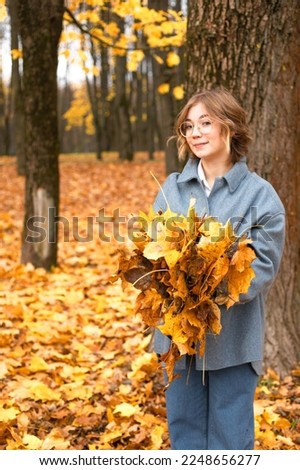 Vertical portrait of stylish, playful, smiling blond woman in eyeglasses holding bouquet of golden oak or maple leaves in hands and looking at camera in park or forest. Exploration, collect herbarium Royalty-Free Stock Photo #2248656277