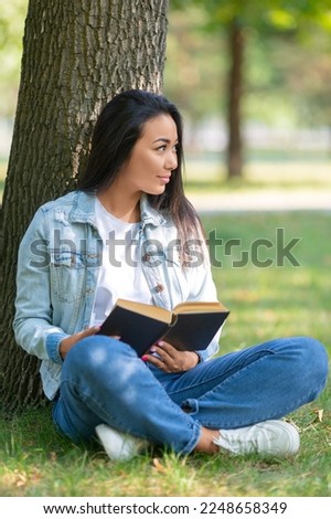 Vertical photo of an asian woman with a book in the park. She sits on the grass in the park and looking by side. The concept of rest from gadgets, smartphones and the Internet. Cropped photo.