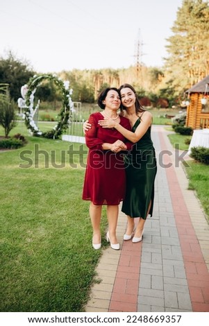 Beautiful smiling girl with her mother hugging. Mother and daughter are walking on green grass at a summer party. Mother's Day. Emerald summer dress. A warm summer evening with the family