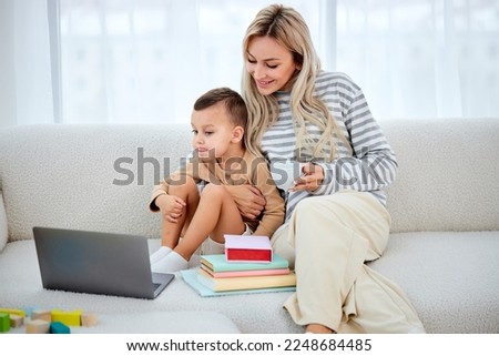Smiling mother with cute lovely son using laptop watching cartoons. happy mum and preschool child sitting on sofa at home, watching video online, mom hugging son, drinking tea. in bright cozy room