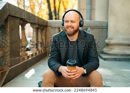 Young bald man listens to a podcast while enjoying a cup of coffee outdoors sitting on some stairs.