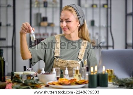 Woman going to mix ingredients to make perfume with many bottles of essential oil. sit behind table using laptop, indoors in cozy room alone. blonde female perfumer creates fragrance Royalty-Free Stock Photo #2248696503