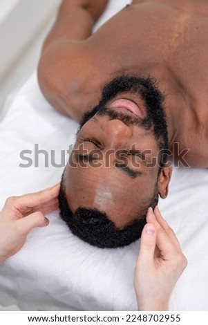 Spa therapist making relaxing massage for handsome middle aged black man. Peaceful bearded man getting healing head massage at modern luxury spa. handsome guy lying on bed couch relaxed, top view