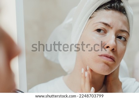 After the shower, the body and head wrapped in a towel. A young woman looks at her face in the mirror and thinks about how to take care of her facial skin. Pimples and acne.