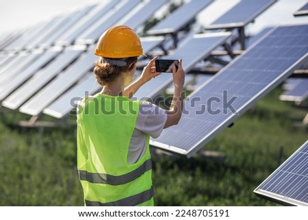 Attractive woman ecological engineer with safety equipment at photovoltaic solar farm taking pictures of solar panels