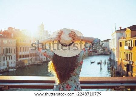 Seen from behind young woman in floral dress with hat on Accademia bridge in Venice, Italy. Royalty-Free Stock Photo #2248710779