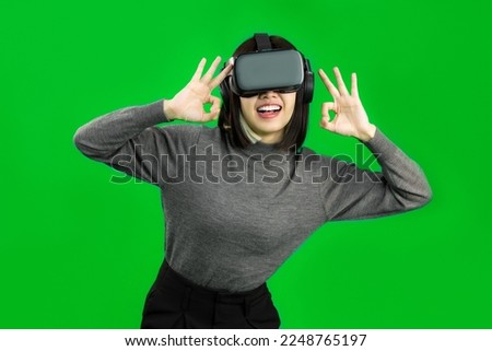 Metaverse. Asian woman in grey long sleeve t-shirt wear vr goggles headset posing ok hands sign, smiling, watching, playing, touching on green screen background. 