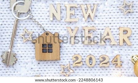 House key with keychain cottage on festive knitted background with stars, lights of garlands. Happy New Year 2024-wooden letters, greeting card. Purchase, construction, relocation, mortgage