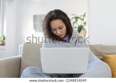 Asian woman wrapped in a cozy blanket sitting on the sofa and working on her laptop