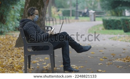 Man with protective mask work at laptop, look at phone in park, sad autumn,