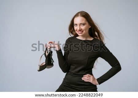 Emotional woman holding high heel shoes in her hands and smiling sweetly in black dress. With light make-up with flowing hair on a light gray background in the studio