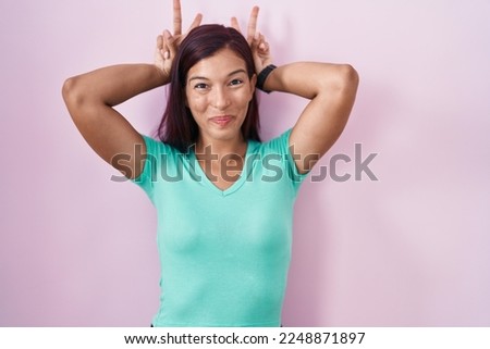 Young hispanic woman standing over pink background posing funny and crazy with fingers on head as bunny ears, smiling cheerful 